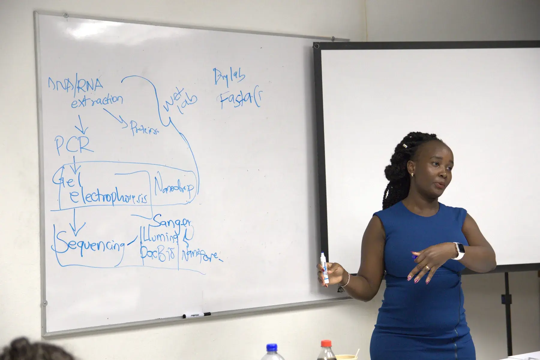 Ruth Nanjala teaching beside a whiteboard with molecular biology notes